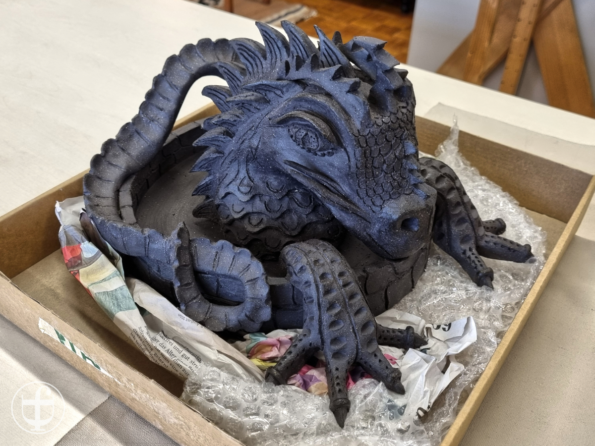 Der fertige Drache Grá sitzt in einem Karton, er hat einen leichten Glanz und ein wunderschönes Blau-Schwarz bekommen vom Brennen von Daniela Joss