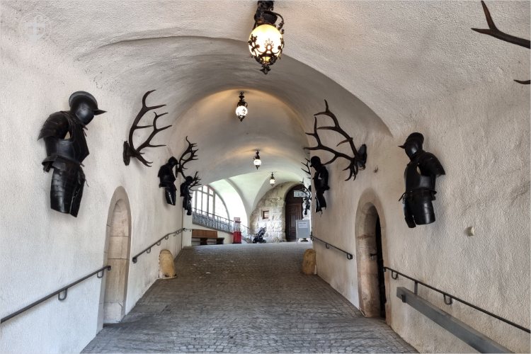 Langer gekalkter Tunnel mit Kopfsteinpflaster. Rüstungen und Hirschgeweihe an den Wänden von Daniela Joss