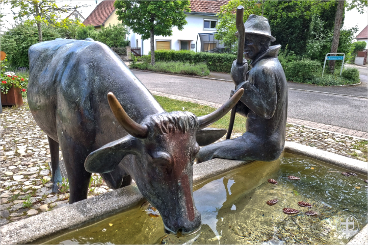Bronzene Skulptur eines Mannes, der mit einem Stab an einem steinernen Brunnen sitzt, während eine Kuh daraus trinkt von Daniela Joss
